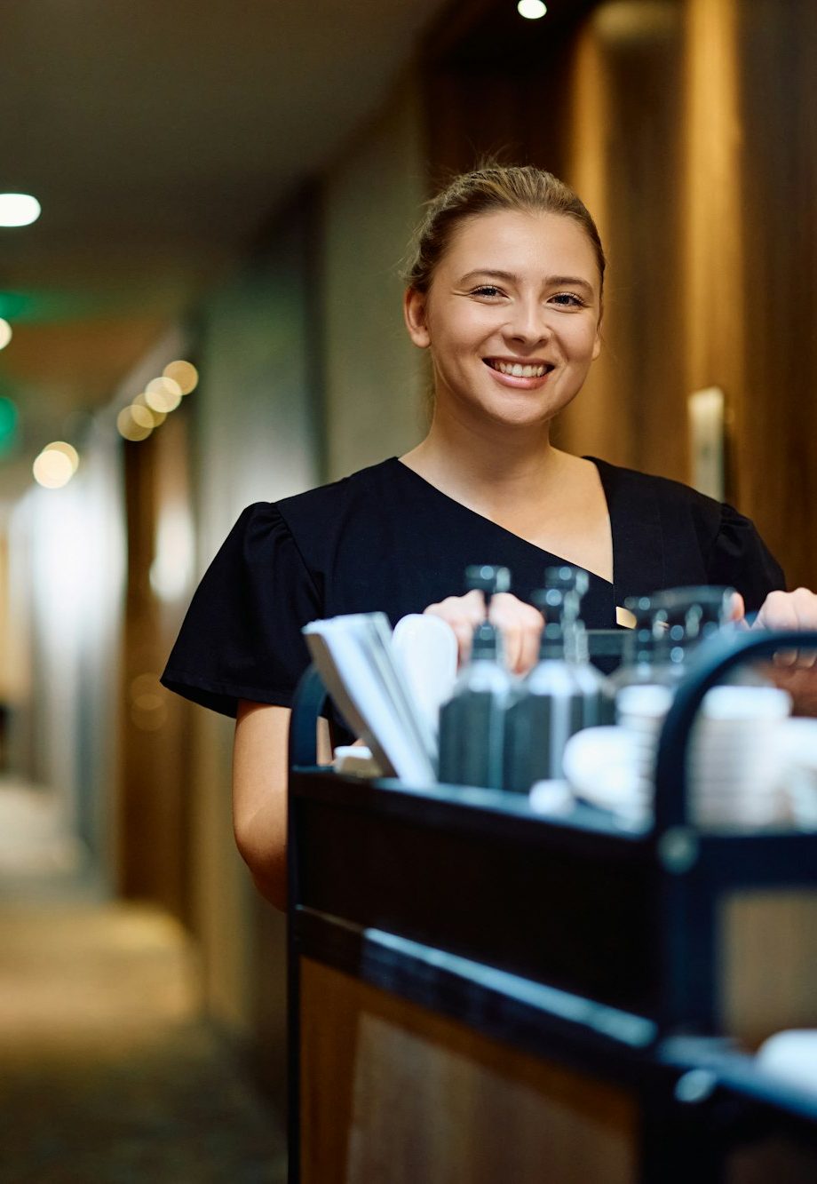 Happy housekeeper with stocked chambermaid's trolley in a hotel looking at camera.